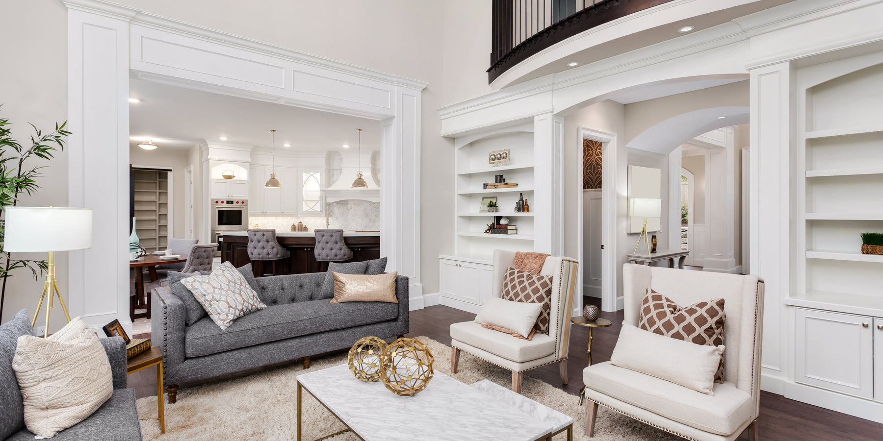 Elegant open-concept living room with modern and classic design elements, featuring a gray sofa, beige armchairs, and a marble coffee table with gold decorative spheres. The space has built-in white bookshelves, a cozy area rug, and a view into a stylish kitchen with white cabinetry, a marble backsplash, and pendant lighting. The room is well-lit with natural light and warm-toned lamps.